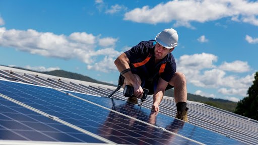 Image of man on rooftop wearing safety gear and installing solar panels on a metal roof.