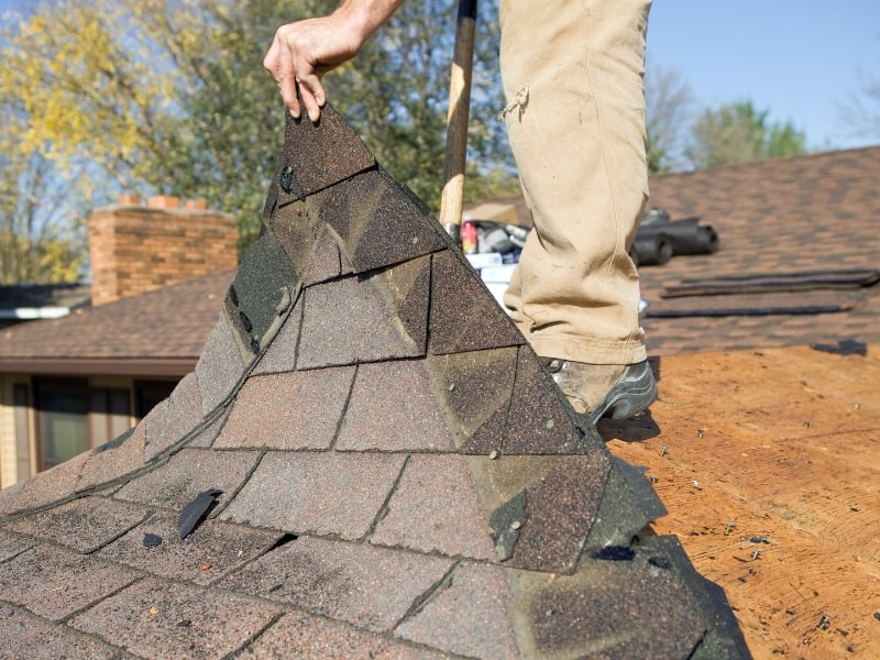 Image of a man in khakis and boots on a roof peeling back and removing old shingles.