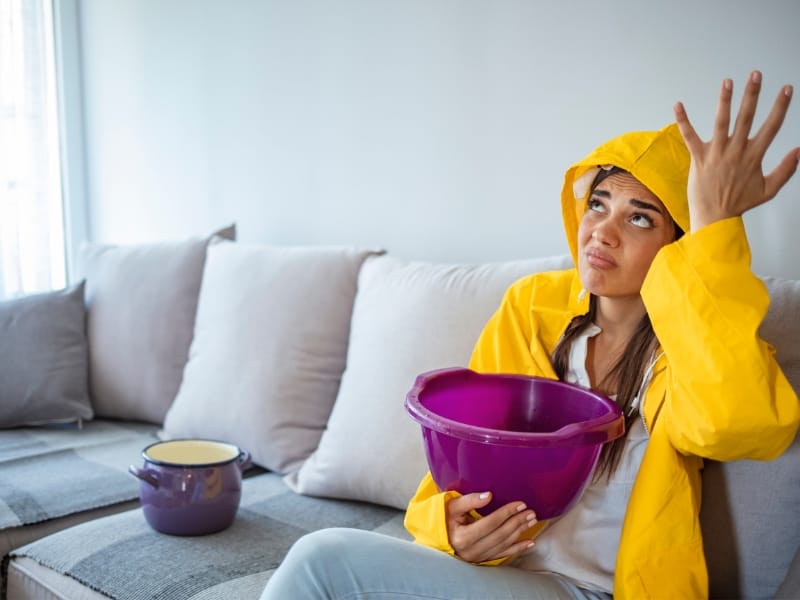 Image of a lady in a yellow raincoat sitting on a beige couch holding a purple bucket to catch water dripping from ceiling.