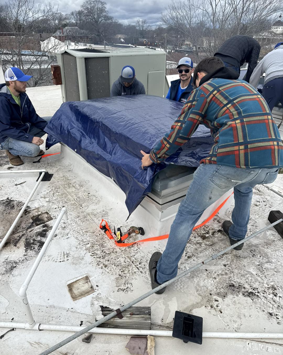 A group of workers from Ridgeline Construction is seen on a storm-damaged commercial roof in Athens, AL, securing a blue tarp over a damaged section. The team is using straps and other materials to prevent further water intrusion. The surrounding area shows storm debris, with the town’s downtown buildings visible in the background under cloudy skies.