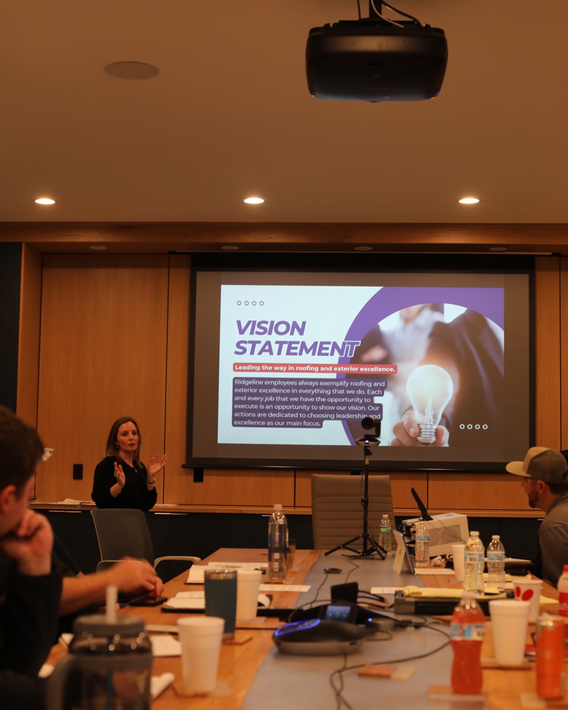 A company meeting at Ridgeline Construction with employees seated around a conference table. Lauren Marsh, the owner, is presenting a slide on the vision statement, which emphasizes leadership in roofing and exterior excellence. The room is well-lit with a projector displaying the presentation.